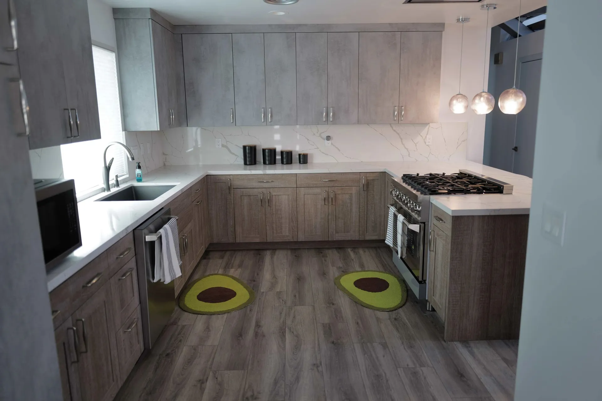 LOT14 HOUSE. Kitchen with gray wood cabinetry, white stone counters, pendant lights, and patterned floor mats.