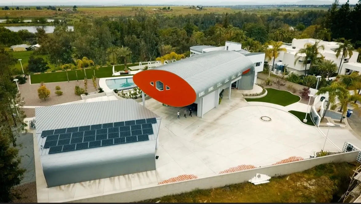 TUBE HOUSE. Aerial view of the curved residential compound with a long silver roof, pool, and planted grounds.